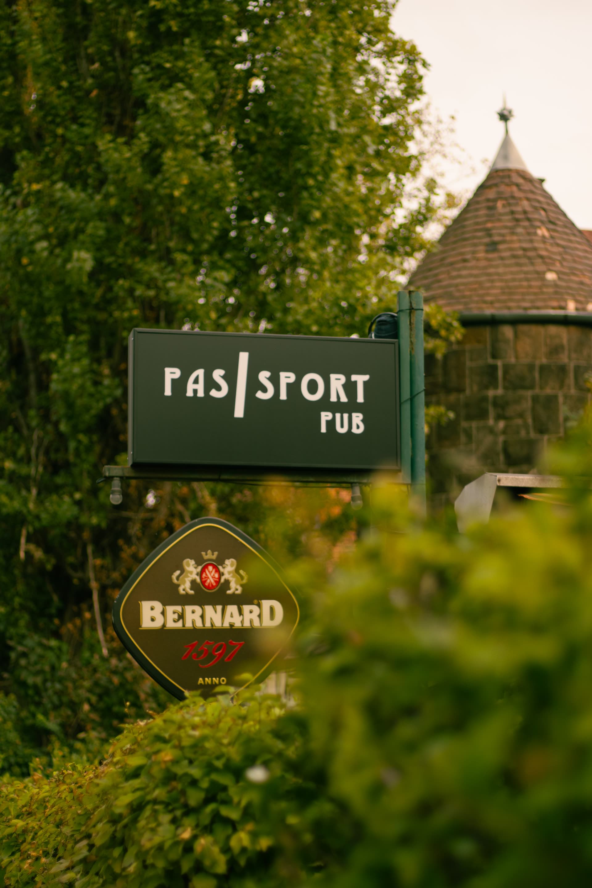 A Passport Pub zöld cégtáblája a fák között Pasaréten, mögötte egy tornyos villa